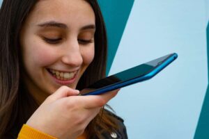 Ragazza sorridente che pigia schermo smartphone ed è intenta a parlare al microfono del device per registrare